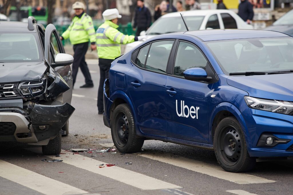 Uber car accident with another vehicle on city street, showing damaged cars and police officers at the crash scene.